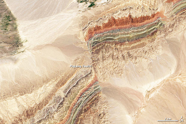 alt: 4) Satelitní snímek zlomu Piqiang v severozápadní Číně. Barevné pásy jsou různé usazené horniny prvohorního stáří.